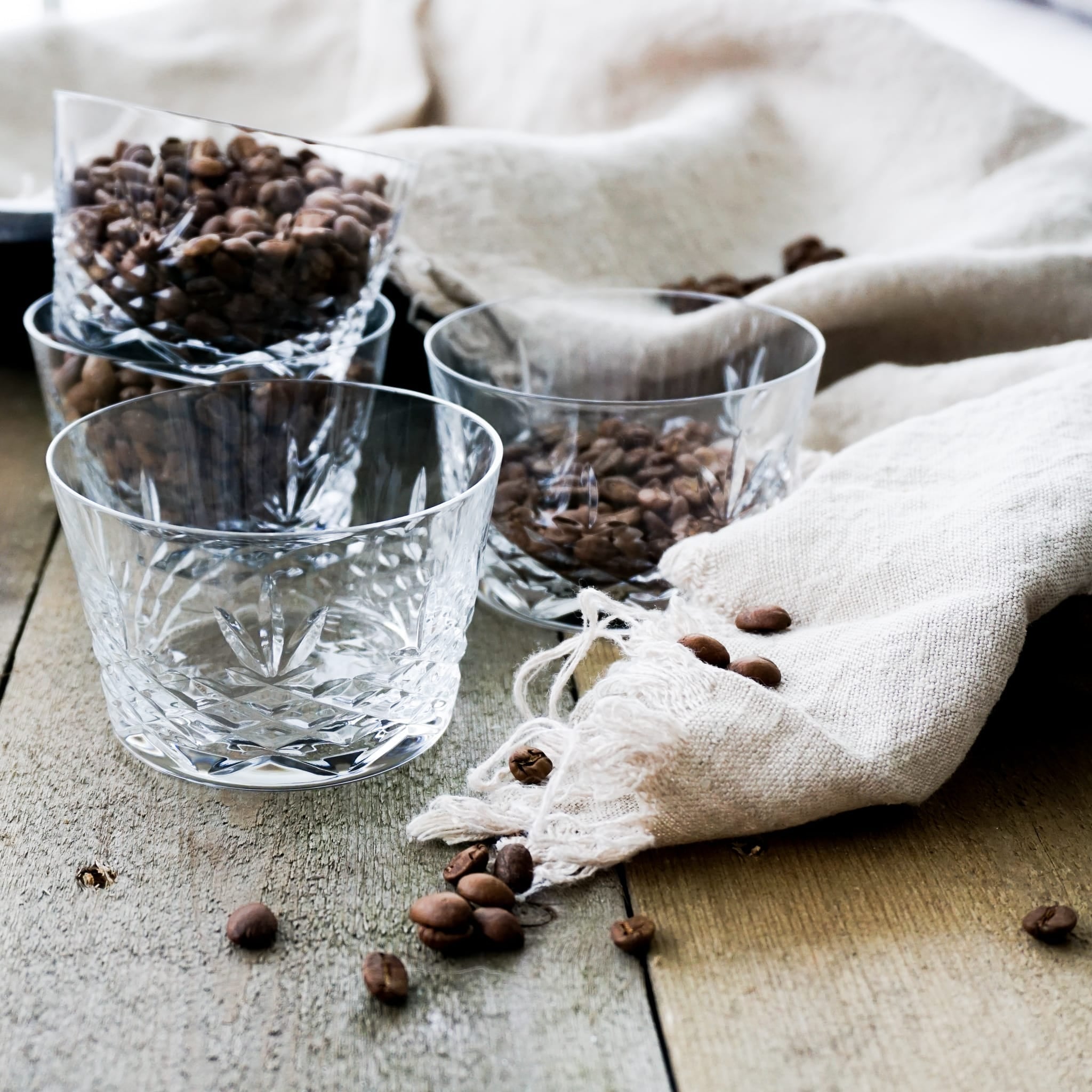Skåler i klart glass med diamantmønster og kaffebønner på rustikk bordflate med tekstil.