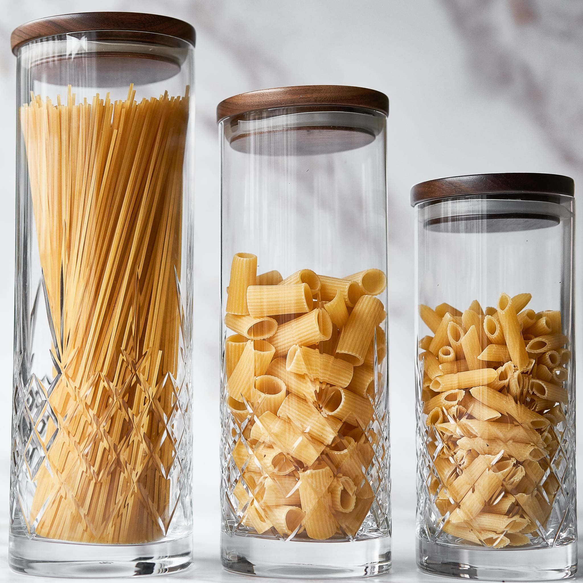 tre klare glasskrukker med rillet skandinavisk design og lokk i tre fylt med tørr pasta