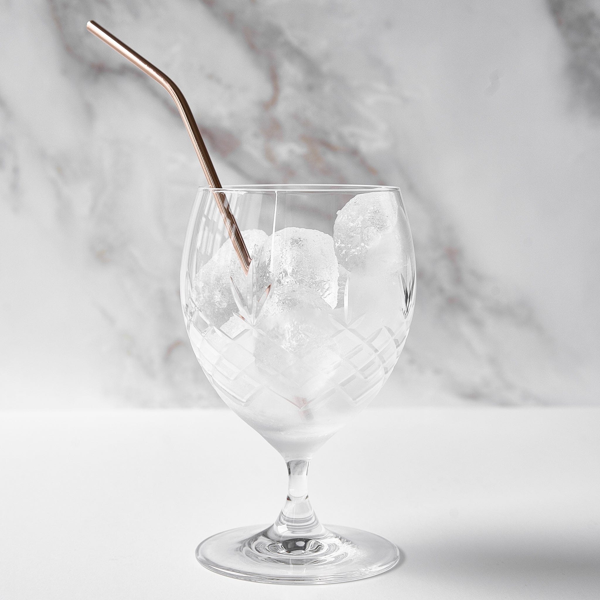 klart glass med diamantmønster fylt med isbiter og metall sugerør på hvit overflate og marmor bakgrunn