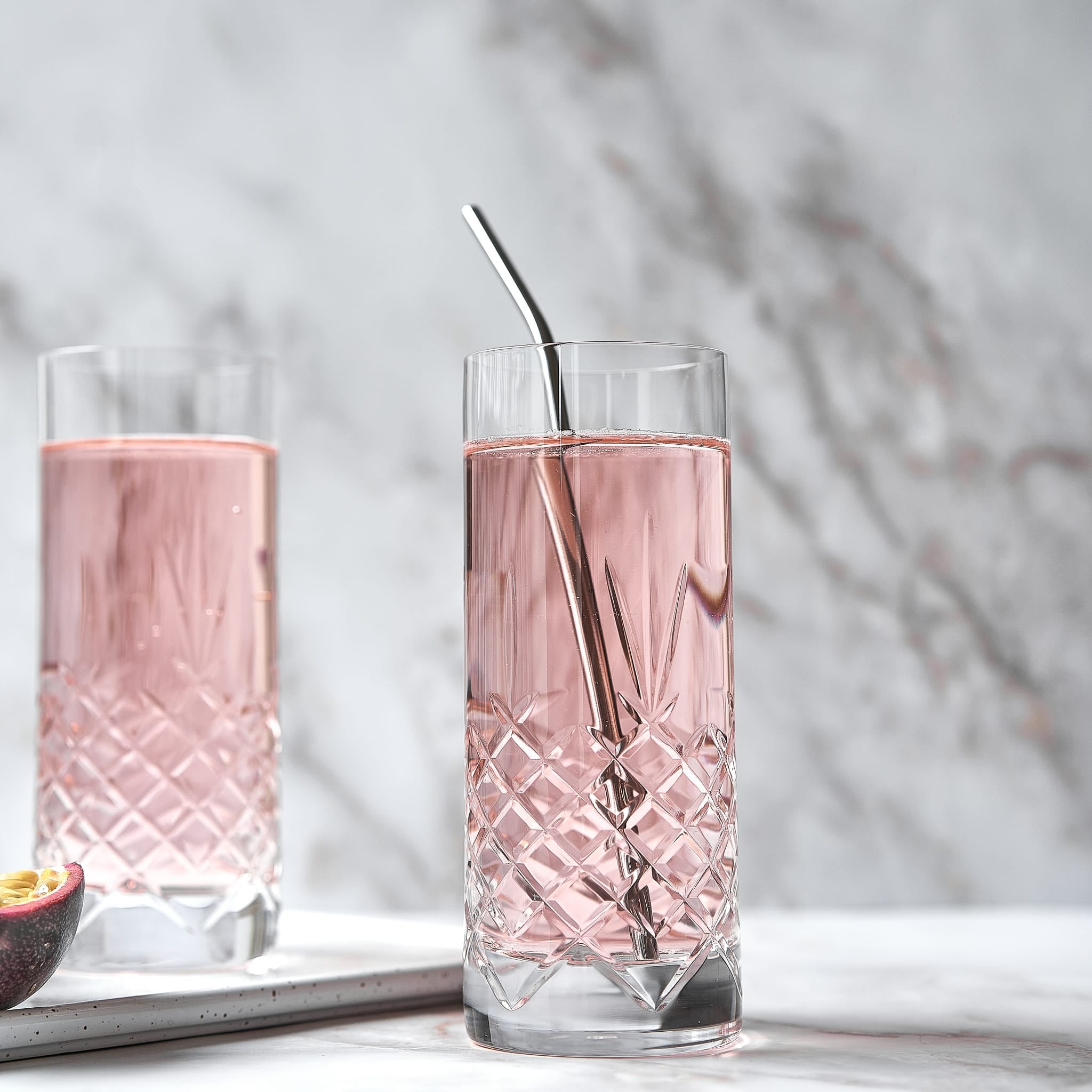 To høye krystallglass med diamantmønster fylt med rosa drikke og rustfri rørepinne på marmorflate.