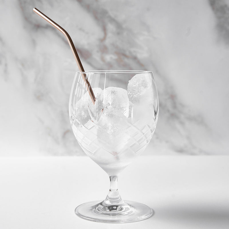 klart glass med diamantmønster fylt med isbiter og metall sugerør på hvit overflate og marmor bakgrunn